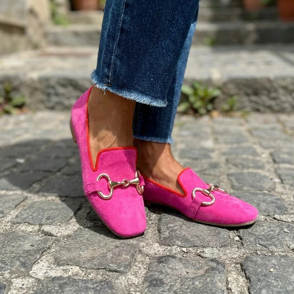 Side view of bright pink loafers with metal detail, paired with jeans on cobblestone surface.