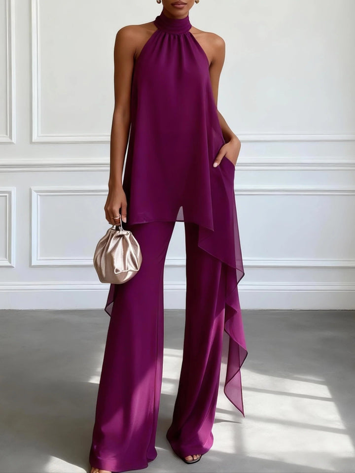 Model wearing a halter neck purple two-piece set with flowing layers and wide-leg pants, holding a small metallic bag in a bright, minimalist room.