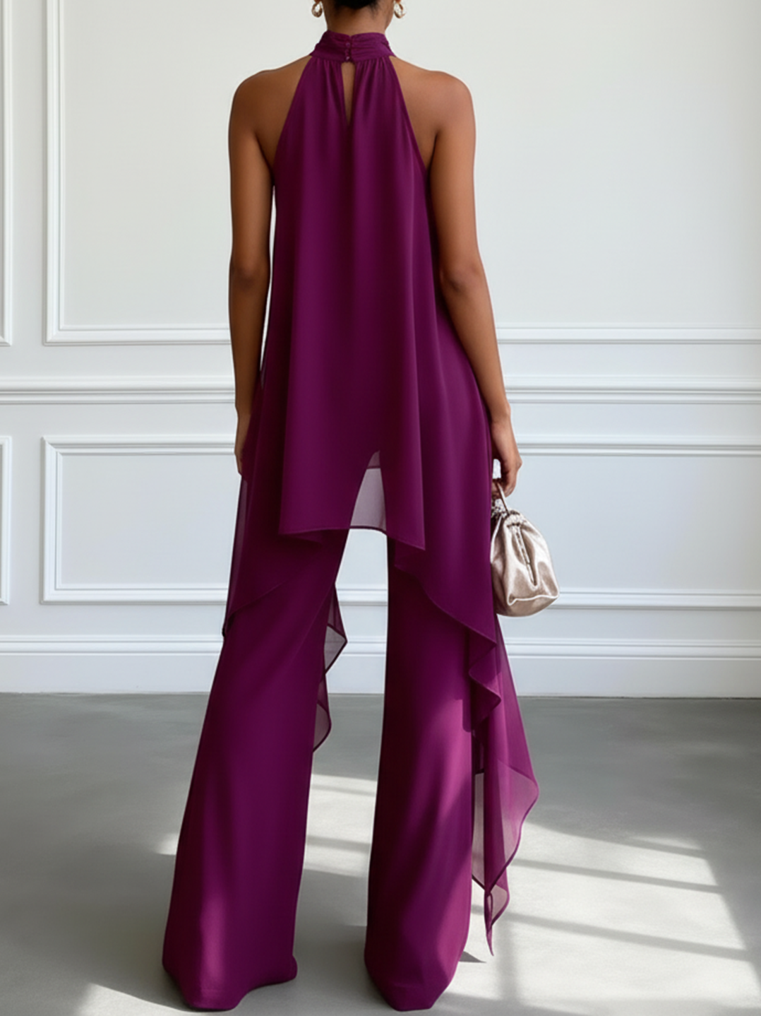 Back view of a model wearing a purple two-piece set with a high neckline and flowing layers, holding a metallic bag, in a bright, minimalist room.