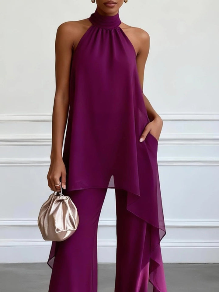 Front view of a model in a purple halter neck two-piece set with flowing layers and wide-leg pants, holding a small metallic bag in a bright room.