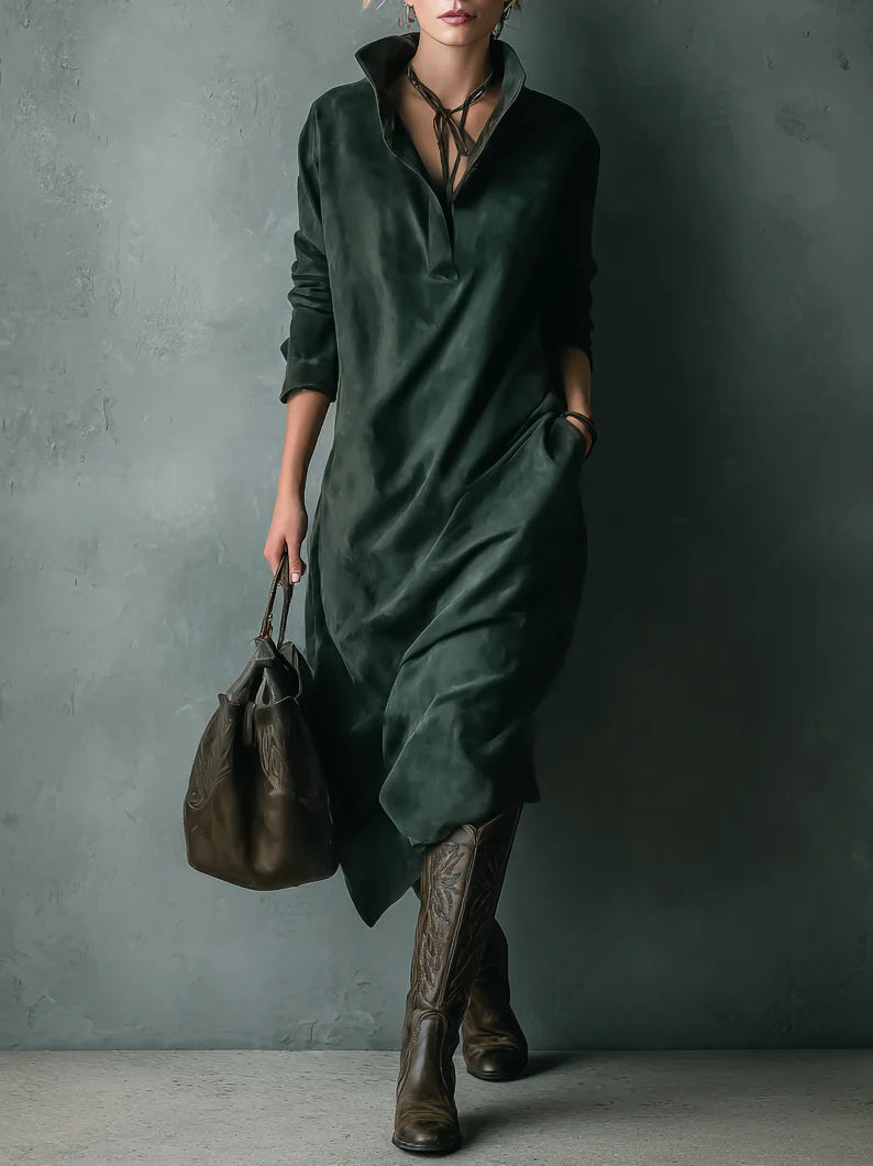 Model in relaxed green velvet dress with pockets, holding a brown bag, wearing tall boots, against textured gray wall.