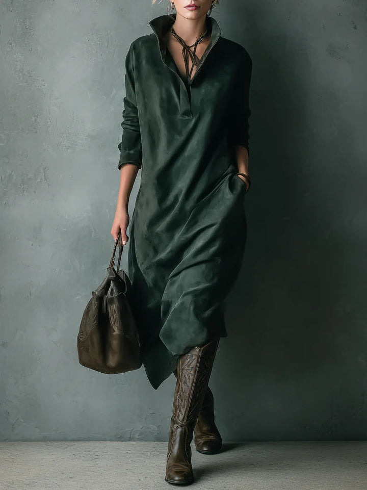 Model in relaxed green velvet dress with pockets, holding a brown bag, wearing tall boots, against textured gray wall.