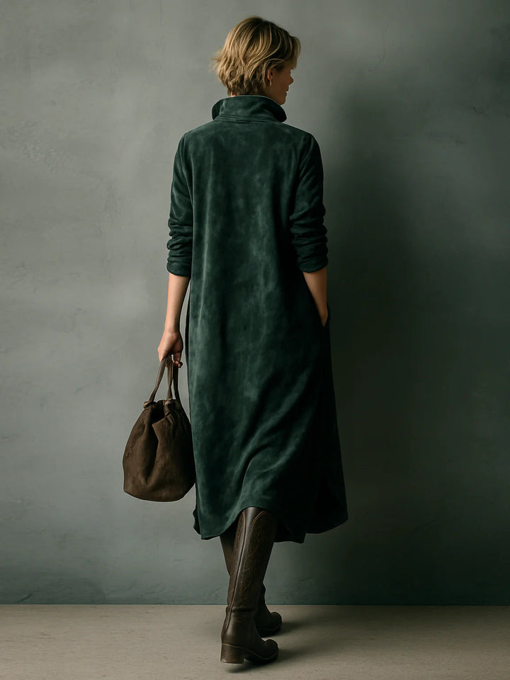 Back view of green velvet dress with pockets, model holding brown bag and wearing tall boots, against gray wall.