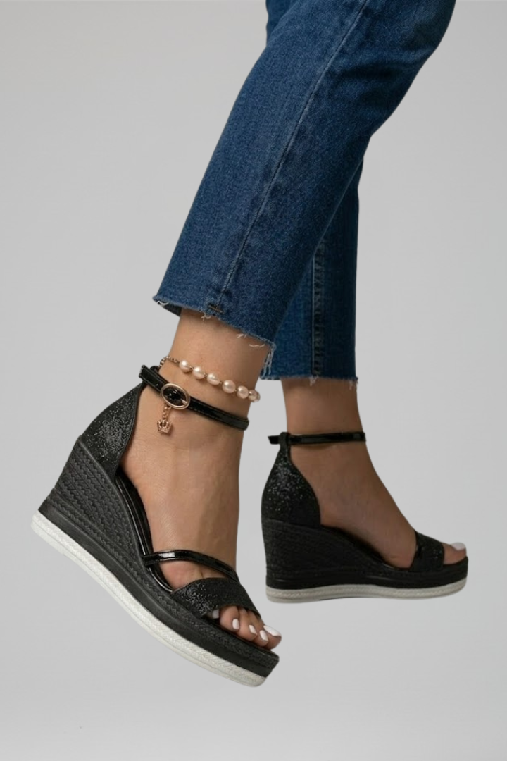 Black cushioned sandals on a raised foot, showing wedge heel and ankle strap in a studio setting.