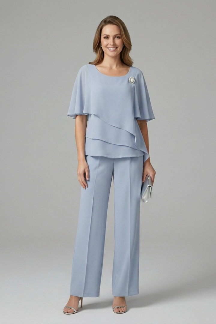 Model wearing light blue layered top and straight-leg trousers set, holding a clutch, in a studio setting.