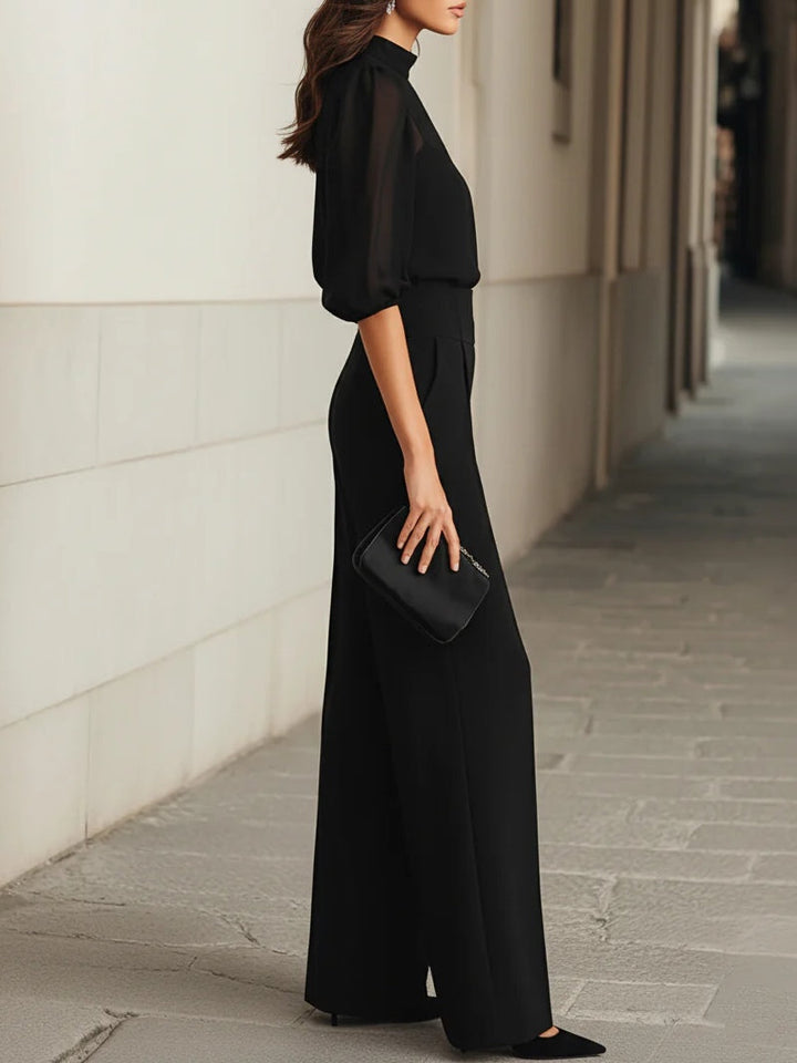 Side view of a sophisticated black three-piece set with a sheer blouse and wide-leg pants, holding a small clutch in an urban scene.