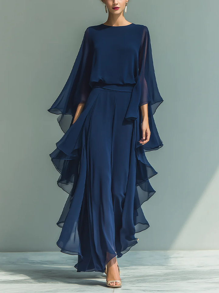 Elegant deep blue two-piece set with flowing layers and cape sleeves, worn with heels in a plain studio setting.