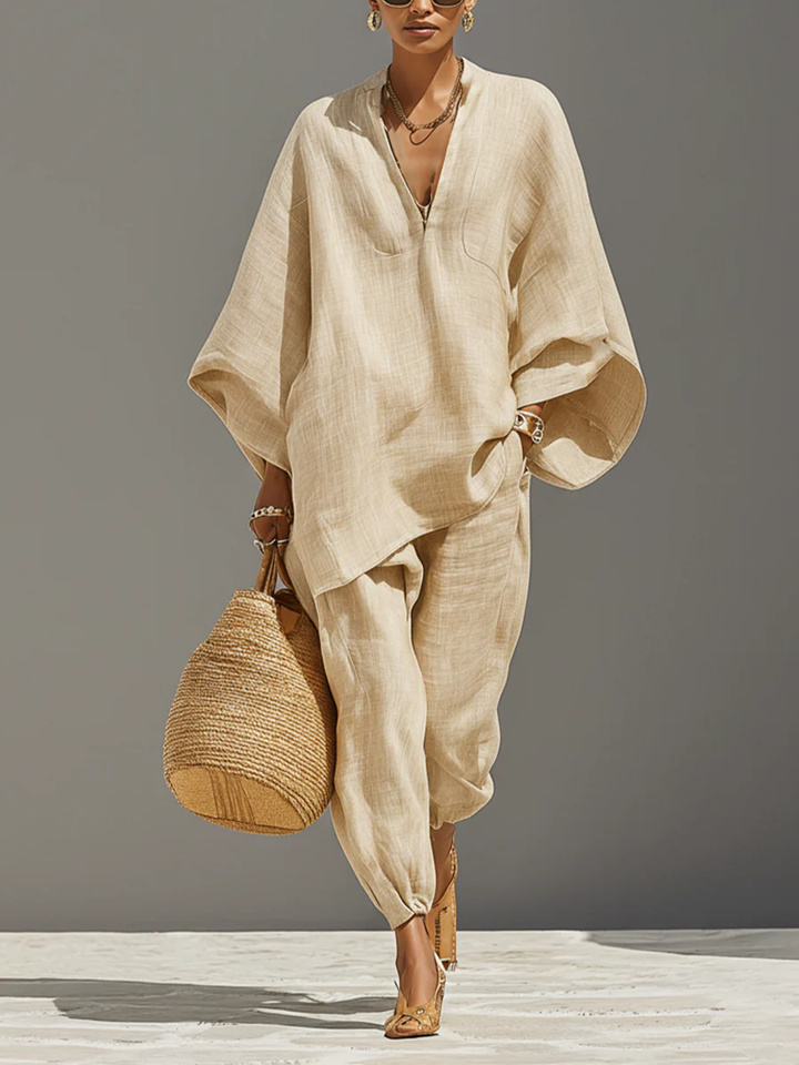 Elegant beige set with flowing top and tapered pants, accessorized with a woven bag, against a neutral backdrop.
