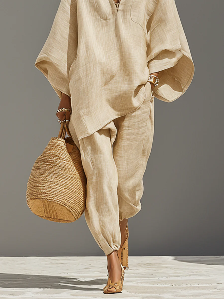 Detailed view of beige set with flowing top and tapered pants, accessorized with a woven bag and heels, on a neutral background.
