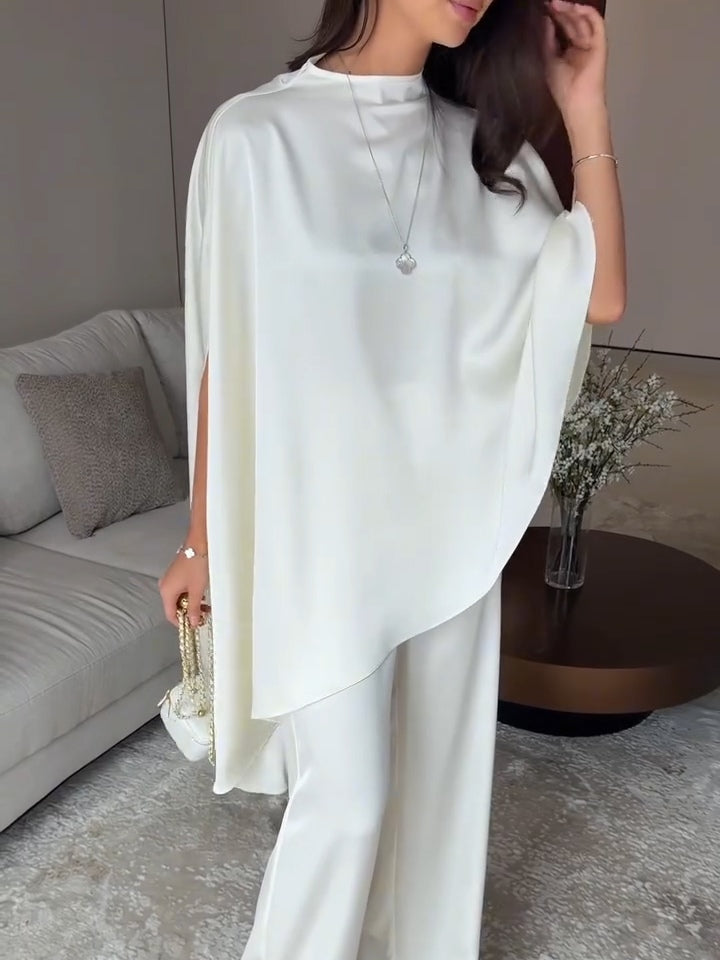 Close-up of woman in white two-piece set with draped design, holding a small handbag in a cozy living room.