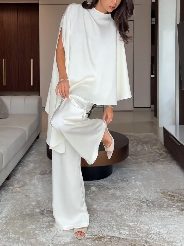Woman adjusting flared pant leg of white two-piece set in a contemporary living room with neutral tones.