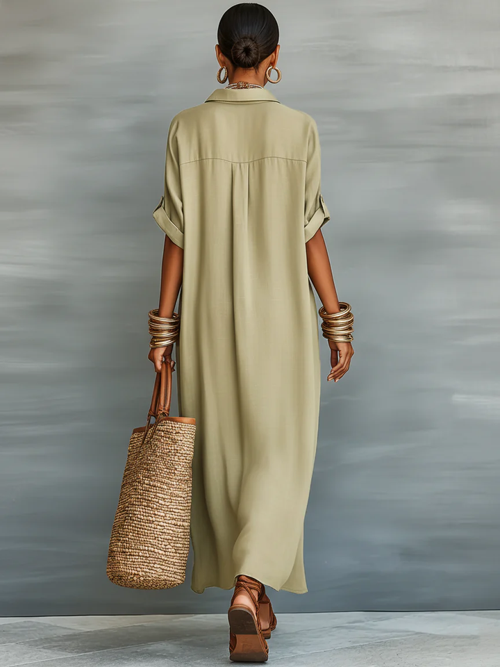 Back view of olive green maxi dress, showing rolled sleeves and relaxed fit. Accessories include hoop earrings, bangles, a woven tote bag, and brown sandals. Studio background.