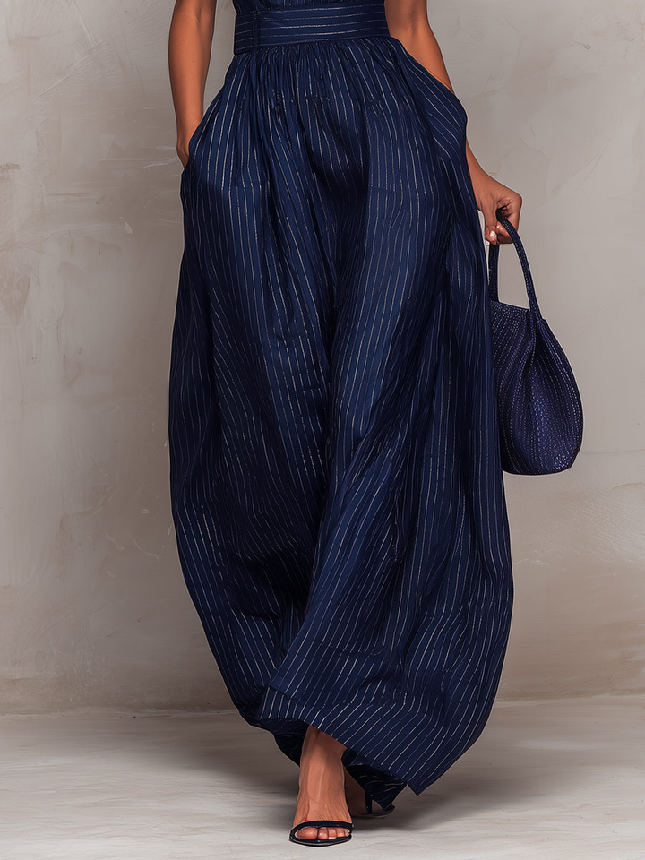 Lower half view of navy wide-leg jumpsuit with vertical stripes, model holding a matching bag, standing on light flooring.