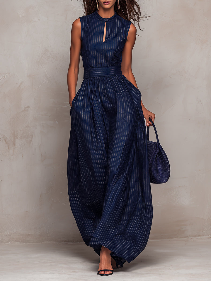 Model in navy wide-leg jumpsuit with vertical stripes, holding a matching bag, in front of a textured beige wall.