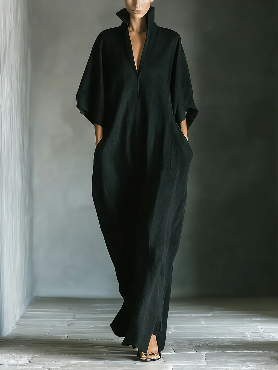 Person wearing a flowing black maxi dress with high collar, standing in a minimalist indoor setting.