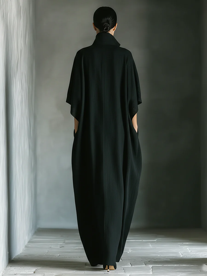 Back view of a black maxi dress with high collar worn by a person in a minimalist indoor setting.