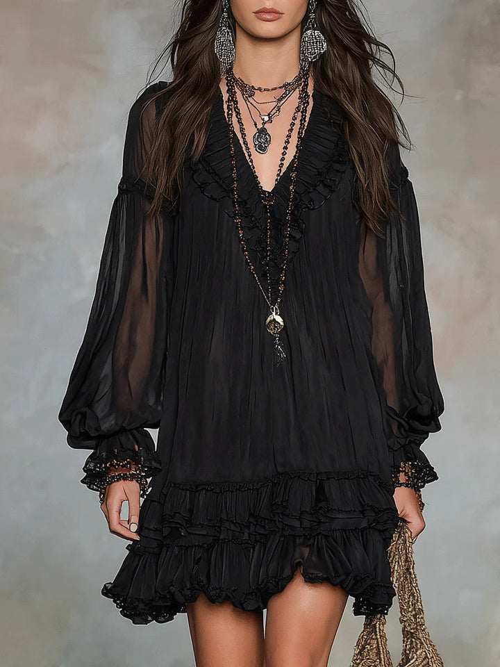 Black mini dress with ruffled neckline and sheer long sleeves, worn in a studio setting with layered necklaces.