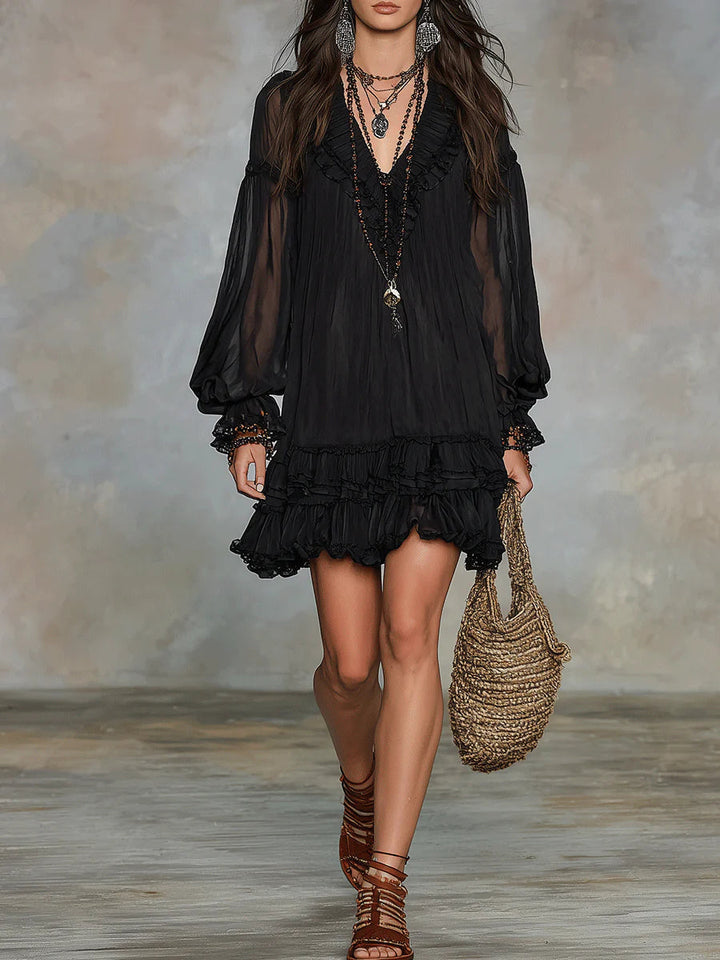 Full view of black mini dress with tiered ruffles and sheer sleeves, accessorized with jewelry in a studio.