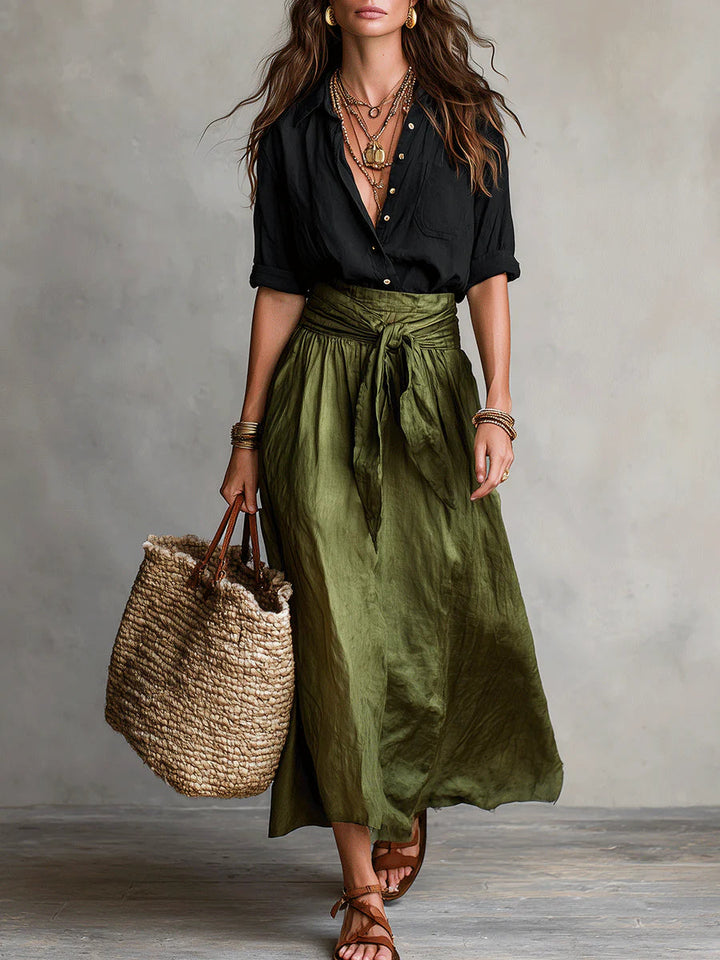Woman in a Vita Two-Piece Set with a black top and green skirt. Holding a woven tote bag. Neutral studio background.