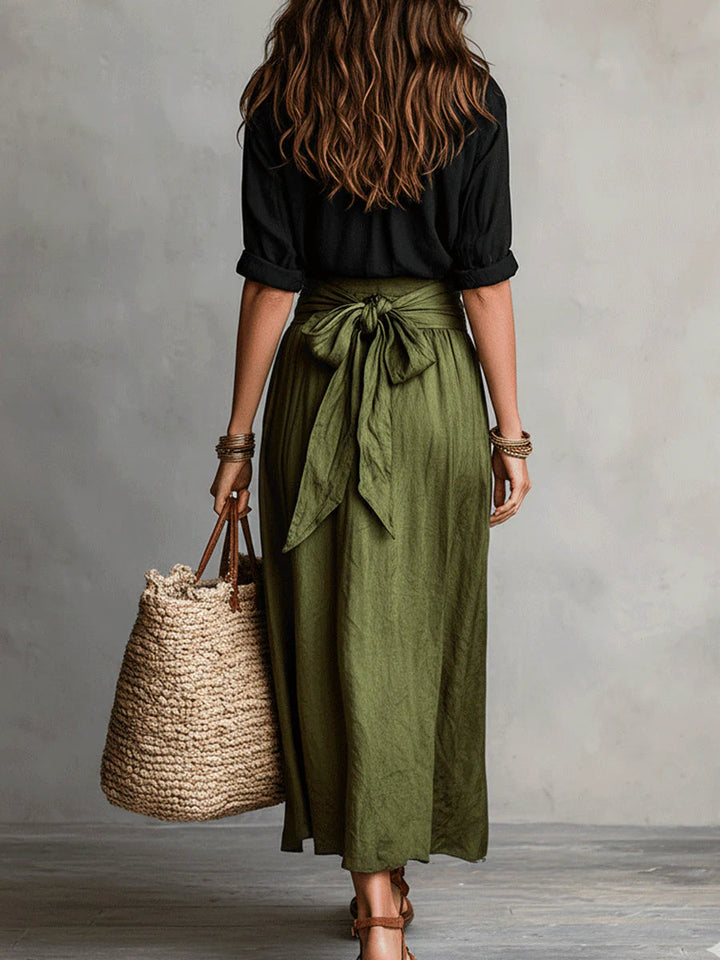 Back view of a woman in a Vita Two-Piece Set, showing tied green skirt and black top. Holding a woven tote bag.