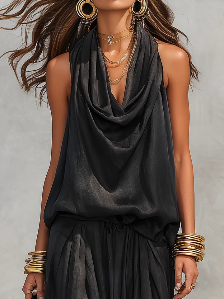 Close-up of woman wearing black two-piece set with draped neckline, large earrings, and bangles, neutral backdrop.