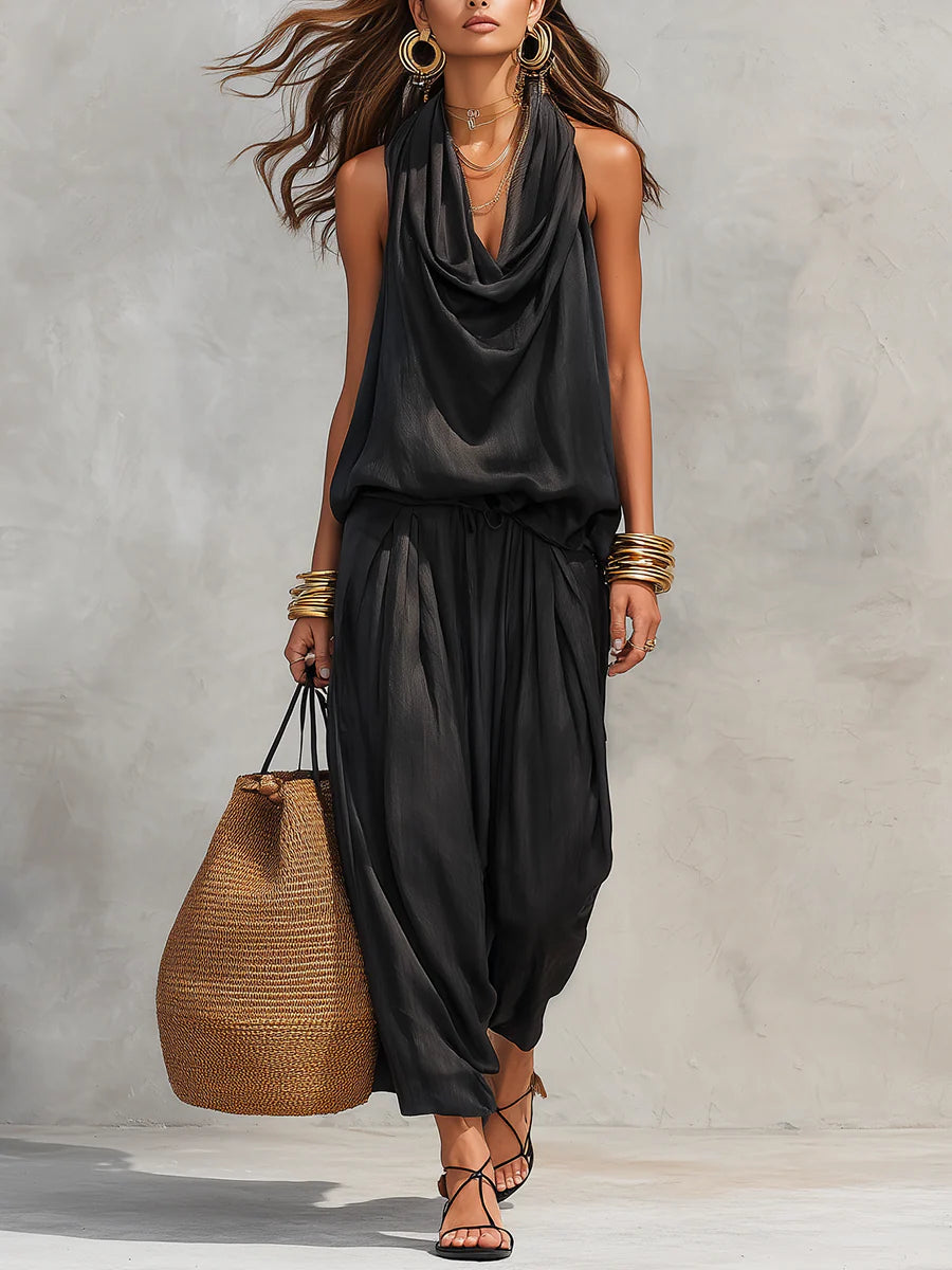 Woman in flowing black two-piece set with draped neckline, holding a straw bag, outdoor neutral background.
