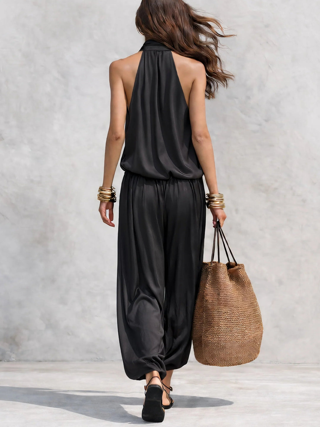 Back view of woman wearing a black two-piece set with halter top, holding a straw bag, outdoor neutral backdrop.