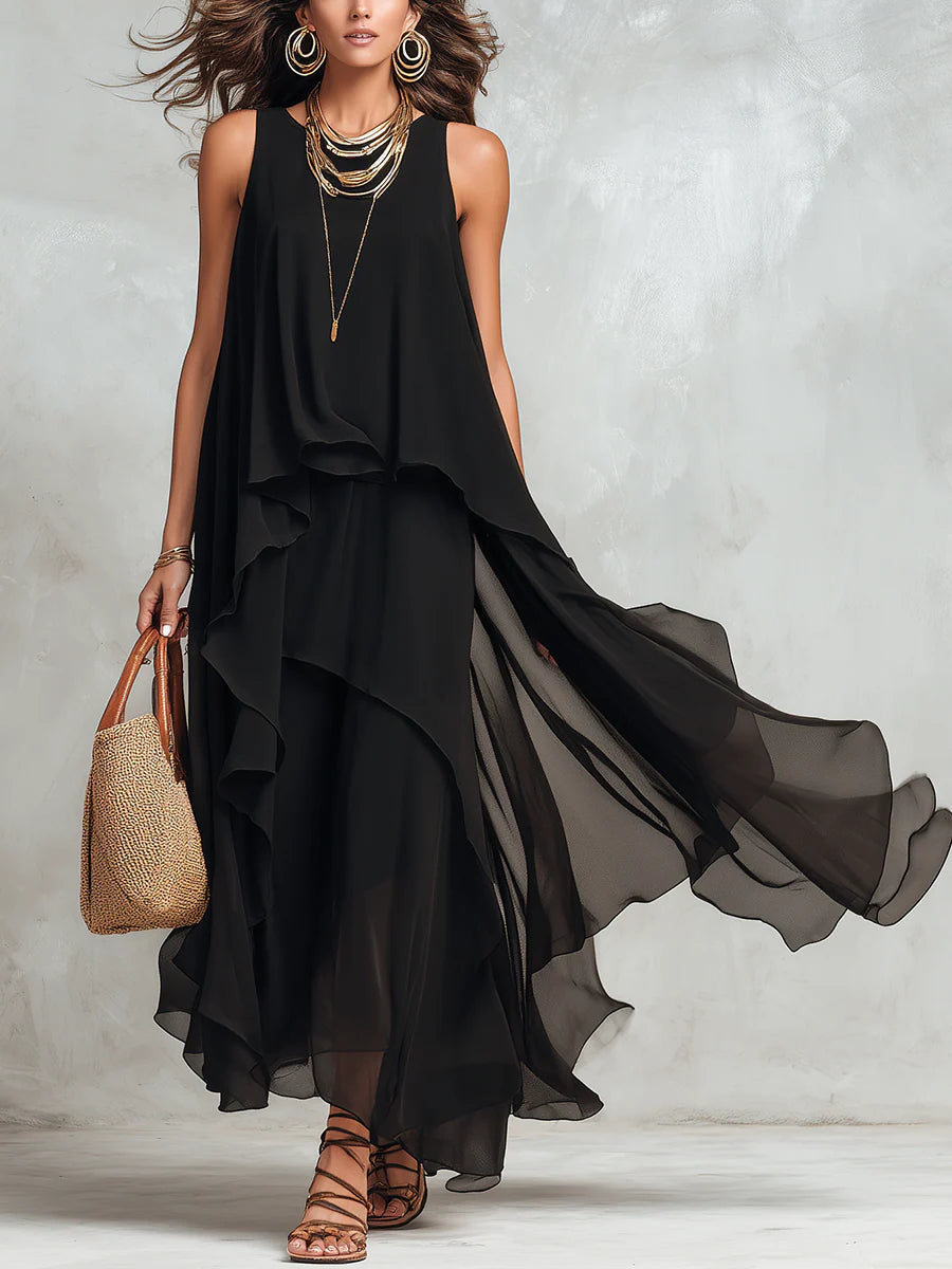 Flowing black maxi dress styled with layered gold jewelry, straw handbag, and brown strappy sandals. Indoor studio background.