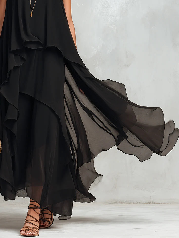 Black maxi dress with flowing layered skirt and brown strappy sandals. Light indoor studio setting.