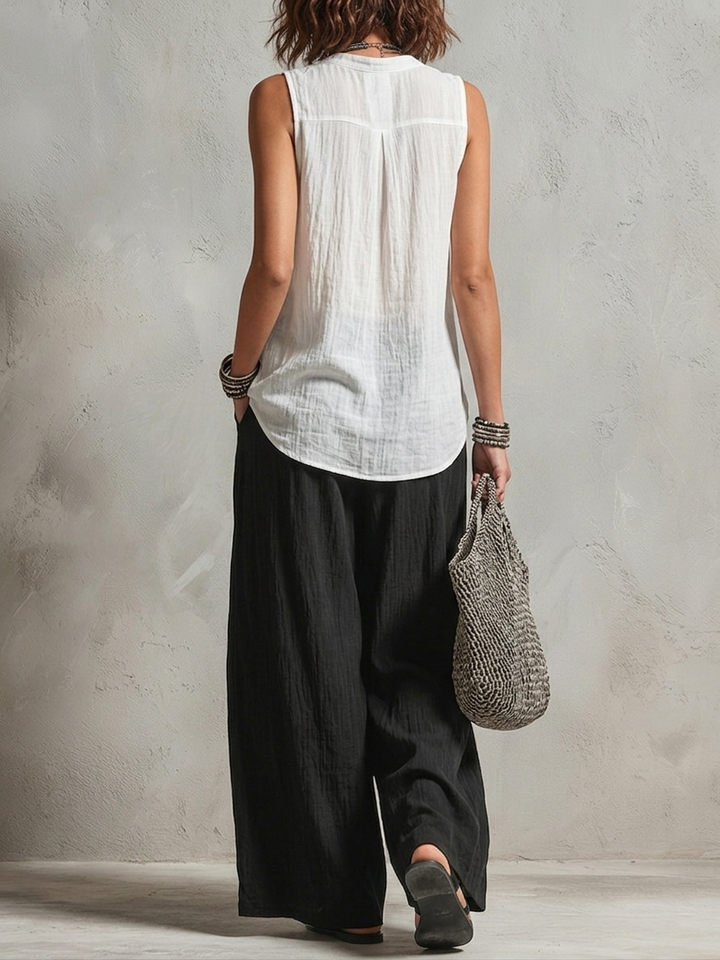 Back view of woman in sleeveless white top and black pants holding a woven bag in a neutral studio.