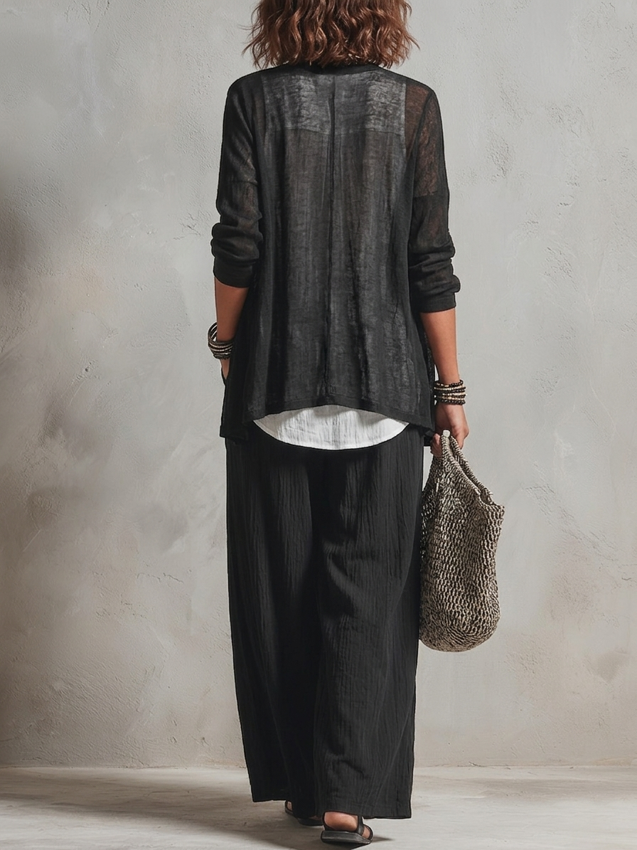 Back view of woman in sheer black top and wide black pants holding a woven bag in a neutral studio.