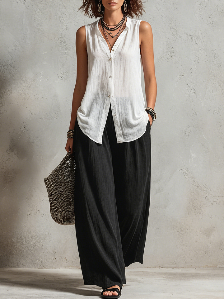 Woman wearing a sleeveless white top and wide black pants with accessories in a minimalist studio setting.
