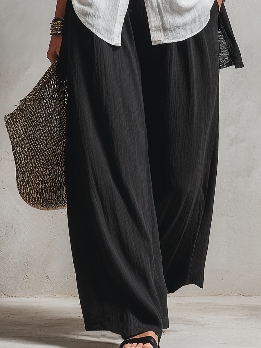 Lower body view of woman in wide black pants and sandals holding a woven bag on a neutral floor.