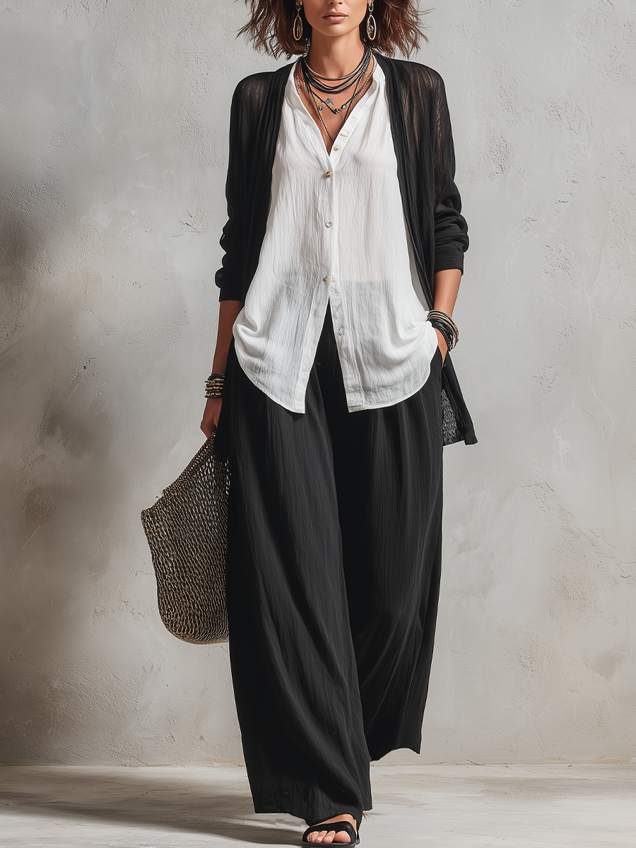 Woman in flowing black and white three-piece set, holding a woven bag, standing in a minimalist studio.