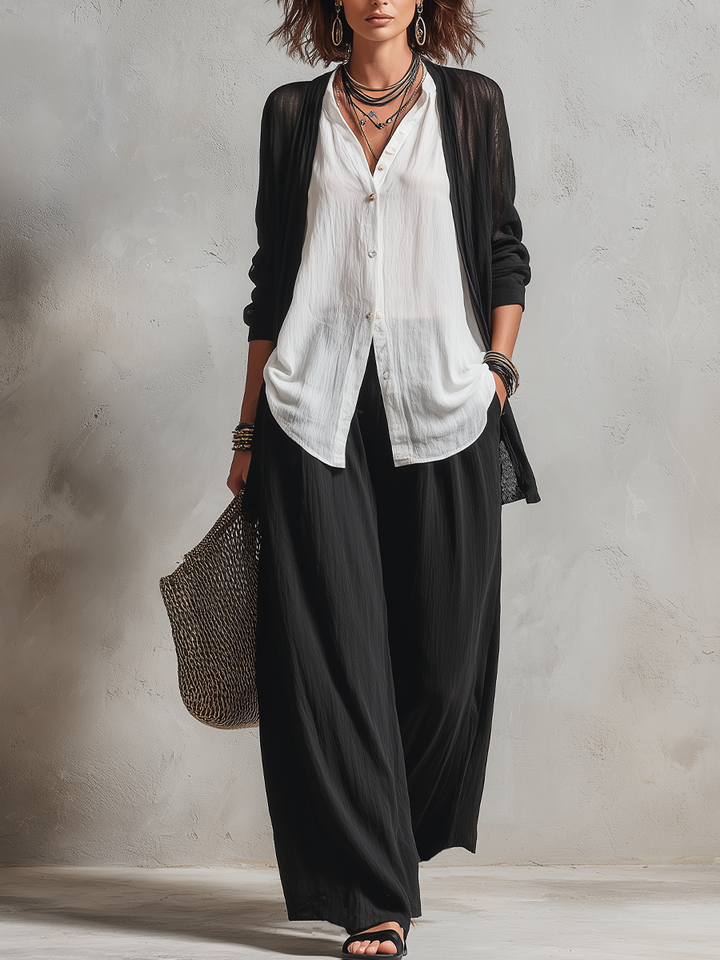 Woman in flowing black and white three-piece set, holding a woven bag, standing in a minimalist studio.
