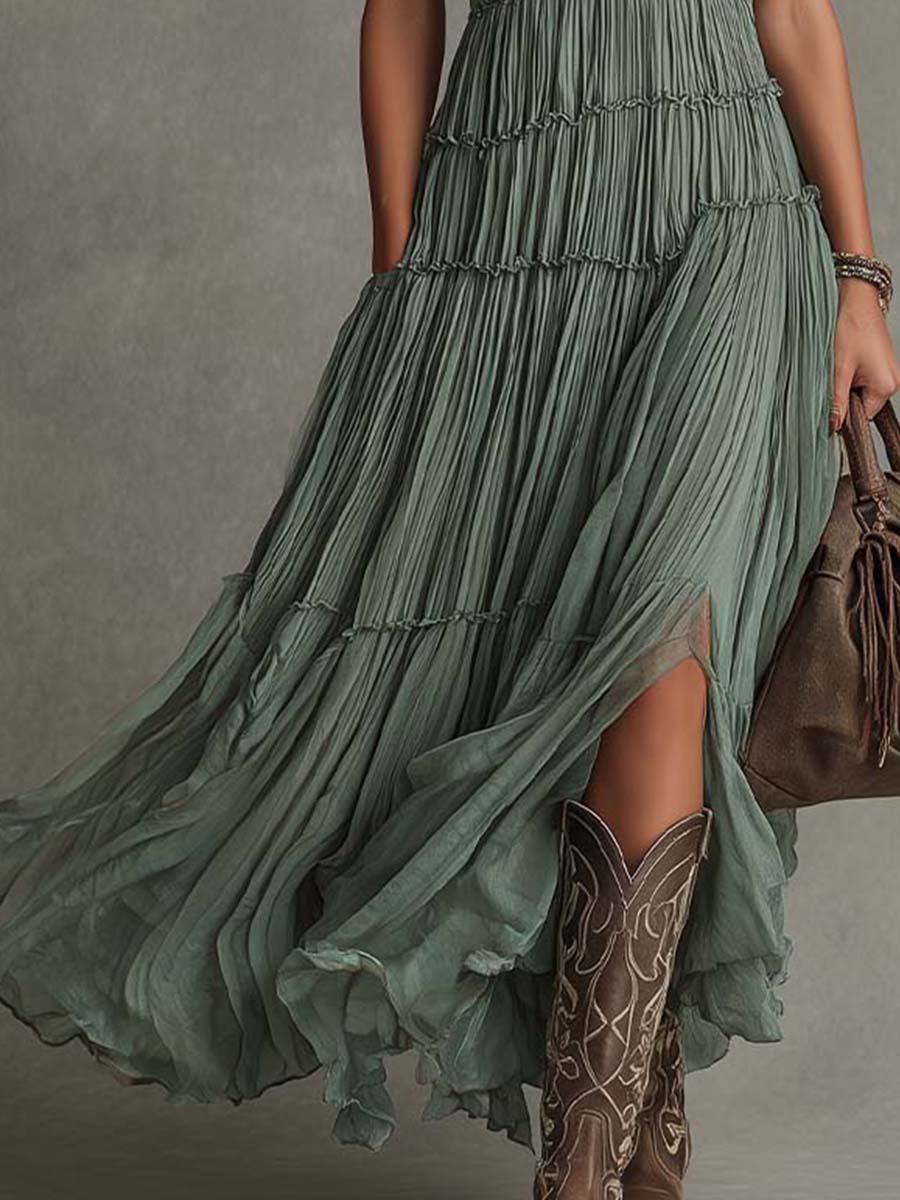 Detail of flowing green maxi dress with cowboy boots, holding a brown bag. Studio background.