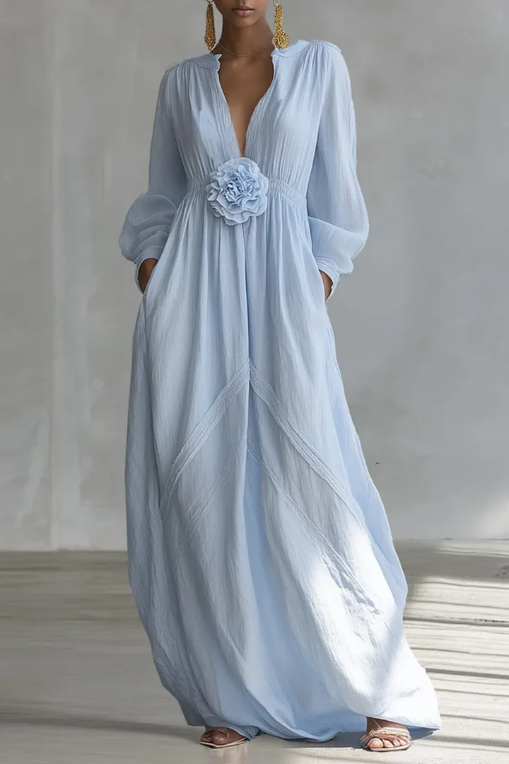 Light blue maxi dress with long sleeves, deep neckline, and floral waist detail, worn in a minimal studio setting.