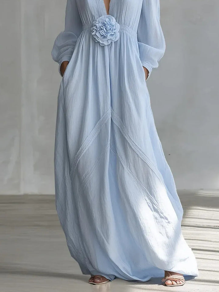 Elegant light blue maxi dress with floral waist detail, pleated design, shown in a minimalist indoor studio setting.