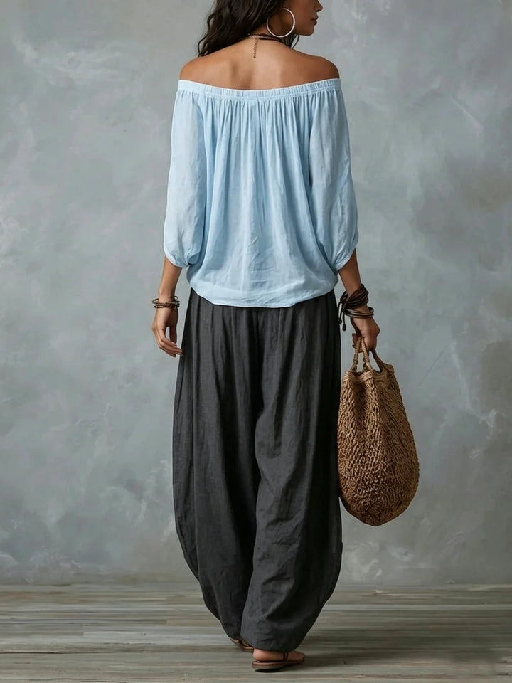 Back view of a woman in a light blue off-shoulder top and black wide-leg pants carrying a woven bag, studio gray backdrop.