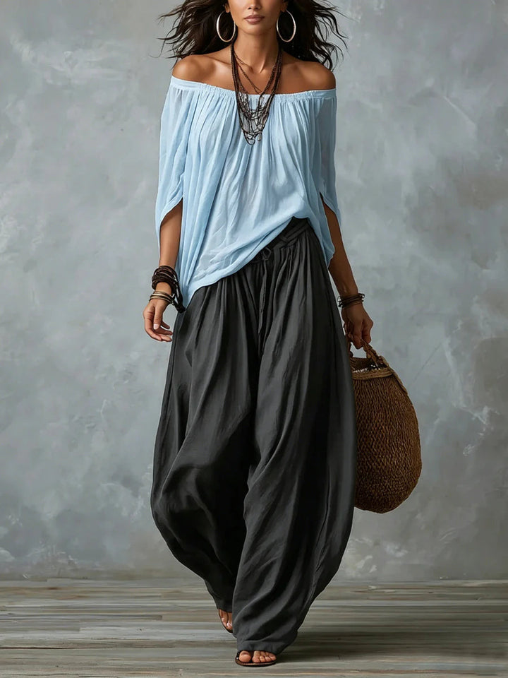 Woman in light blue off-shoulder top and black wide-leg pants holding a woven bag, standing in a studio with a gray backdrop.