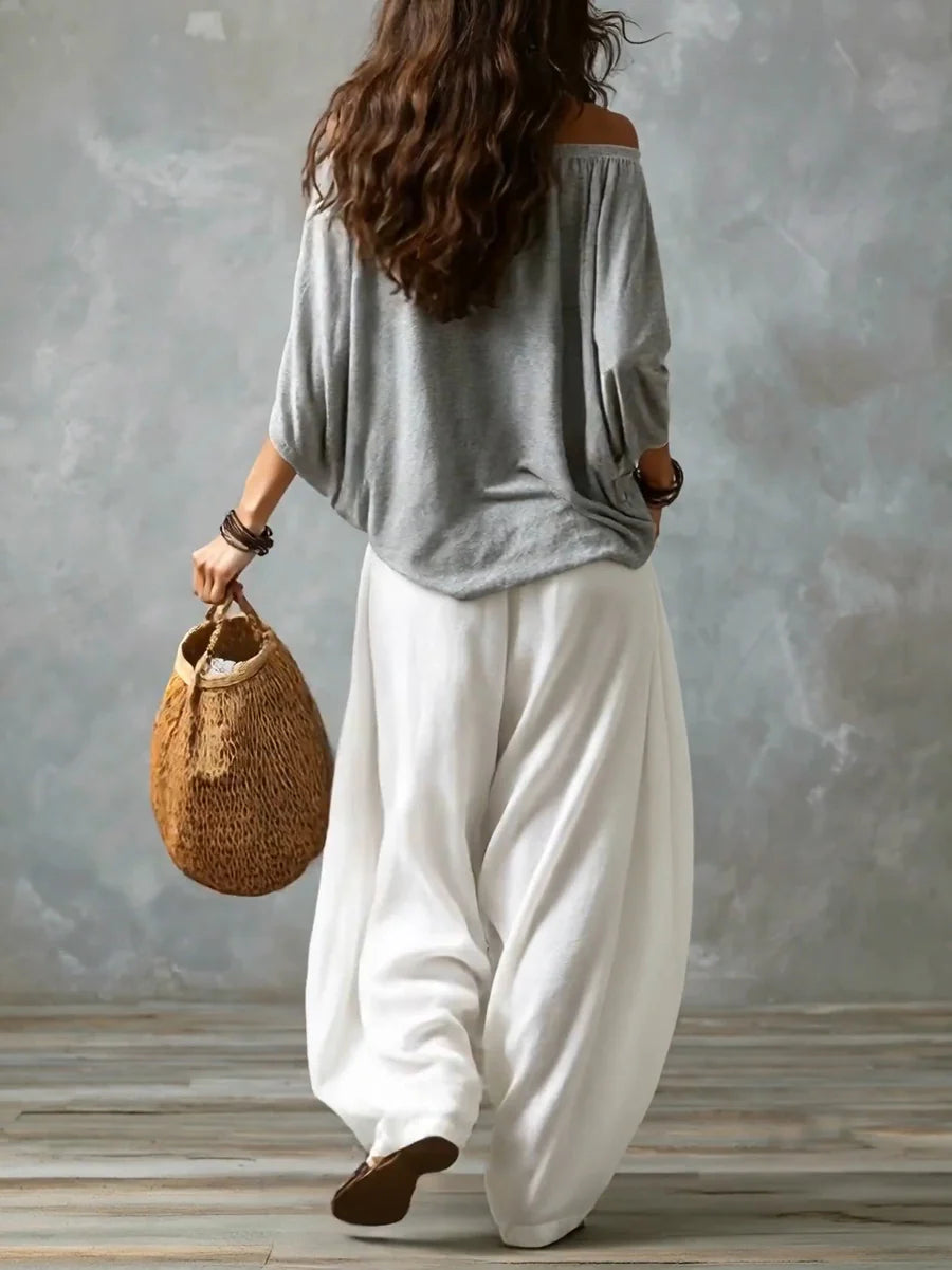 Back view of a woman in a gray off-shoulder top and white wide-leg pants holding a woven bag in a studio with gray backdrop.