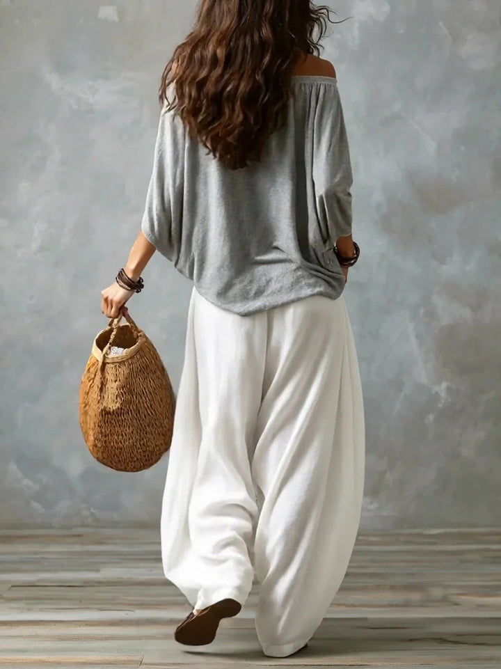 Back view of a woman in a gray off-shoulder top and white wide-leg pants holding a woven bag in a studio with gray backdrop.