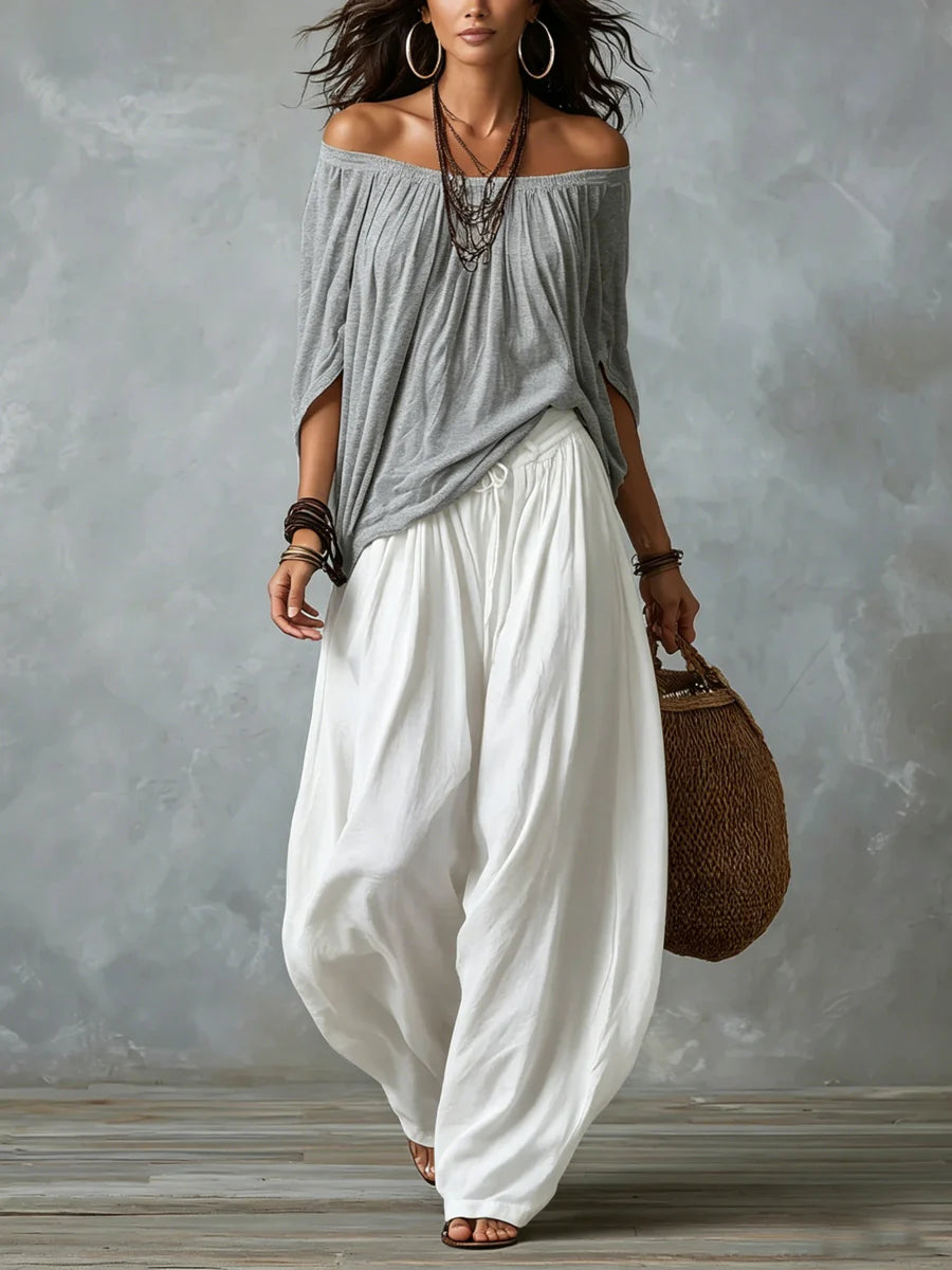 Woman wearing a gray off-shoulder top and white wide-leg pants, holding a woven bag in a studio with a gray textured background.