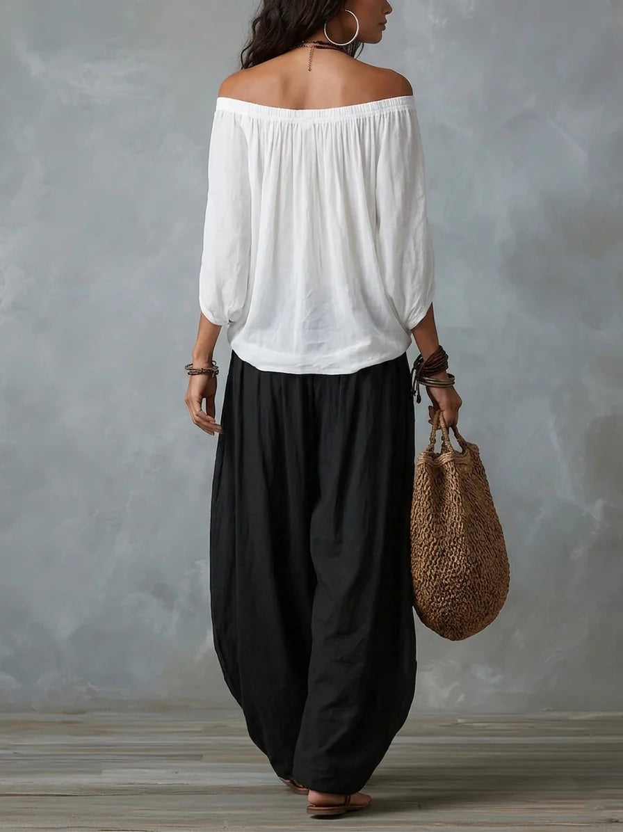 Back view of woman in a white off-shoulder top and black wide-leg pants, carrying a woven bag, studio setting with gray backdrop.