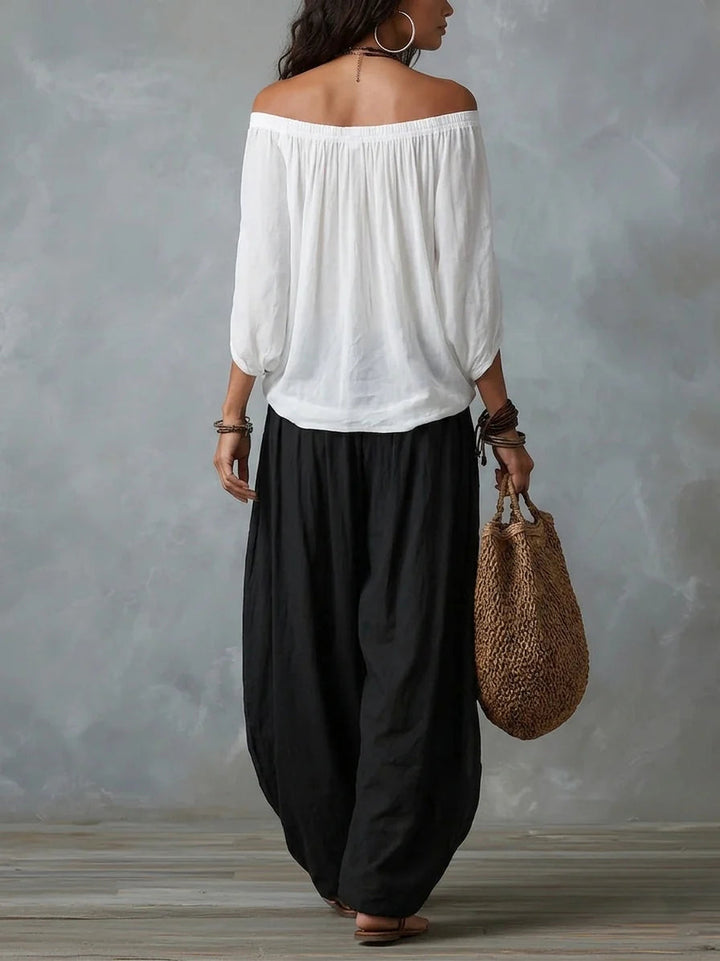 Back view of woman in a white off-shoulder top and black wide-leg pants, carrying a woven bag, studio setting with gray backdrop.