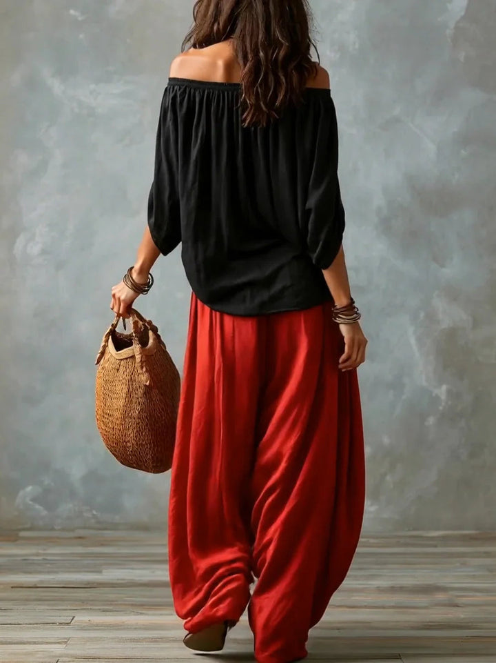 Back view of a woman in a black off-shoulder top and red wide-leg pants carrying a woven bag against a studio gray backdrop.