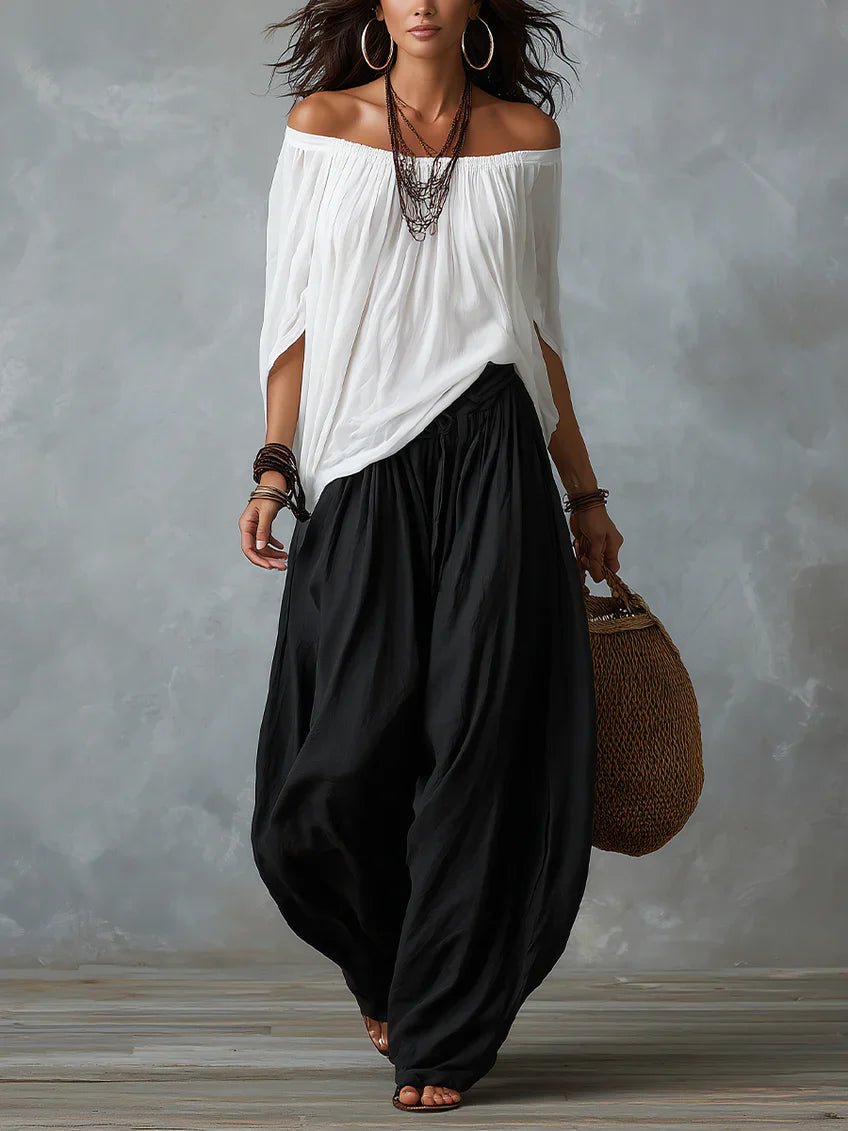 Woman in a white off-shoulder top and black wide-leg pants holding a woven bag, standing in a studio with a textured gray background.