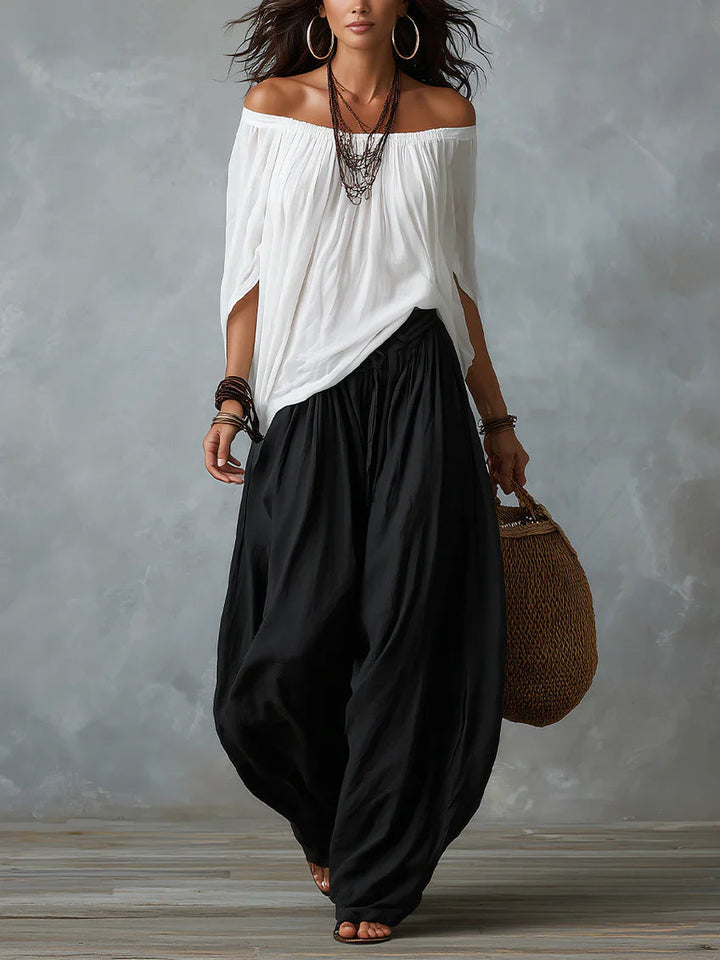 Woman in a white off-shoulder top and black wide-leg pants holding a woven bag, standing in a studio with a textured gray background.