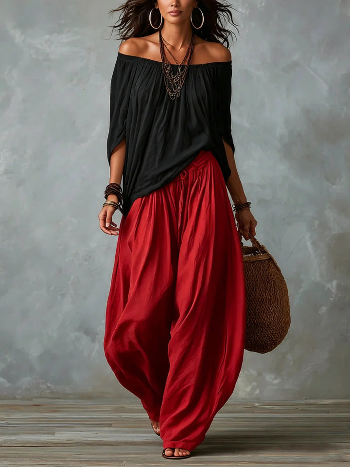 Woman in a black off-shoulder top and red wide-leg pants holds a woven bag in a studio featuring a gray textured background.