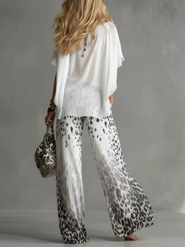 Back view of a woman in a flowing white top and leopard print pants, carrying a matching bag in a studio setting.